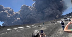 People stand on a rocky landscape taking photos as thick, dark volcanic ash clouds rise dramatically into the sky during an eruption. Cars and buses are visible on the right side.