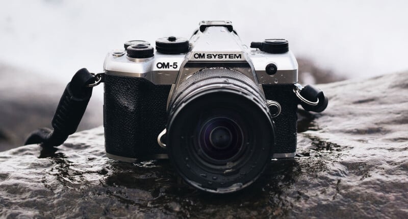 A close-up of a black and silver OM System OM-5 camera with a lens, resting on a wet rock with water droplets visible on its surface. The background is blurred.