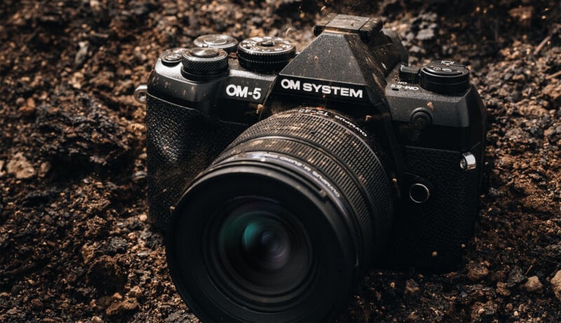 A black OM SYSTEM OM-5 digital camera sits on dark soil, its body and lens dusty, suggesting it has been used in rugged outdoor conditions.