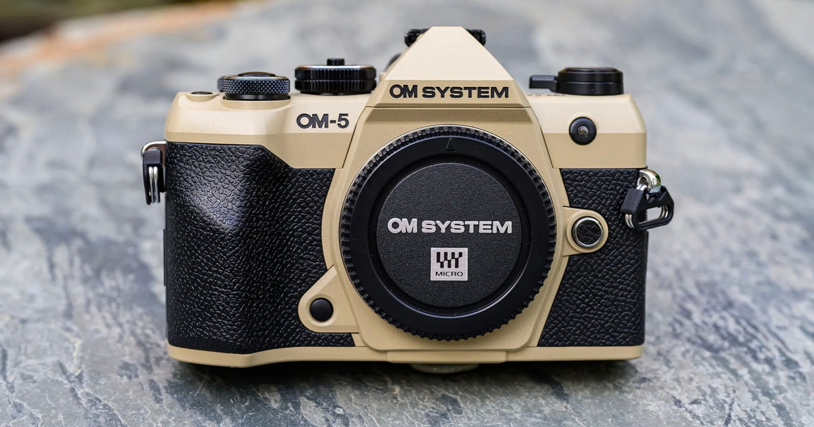 A tan and black OM SYSTEM OM-5 camera sits on a gray stone surface. The camera has a textured grip and a body cap labeled "OM SYSTEM Micro" on the front.