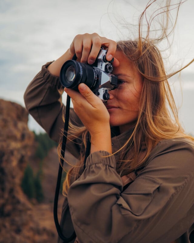 A woman with long hair and a brown jacket is outdoors, holding a vintage camera up to her eye, taking a photo. The wind blows her hair, and there are rocky cliffs and a cloudy sky in the background.