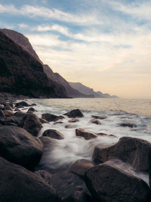 Rocky shoreline with waves gently washing over large boulders, steep cliffs rising along the left, and a cloudy sky above, creating a peaceful and dramatic coastal scene at sunset or sunrise.