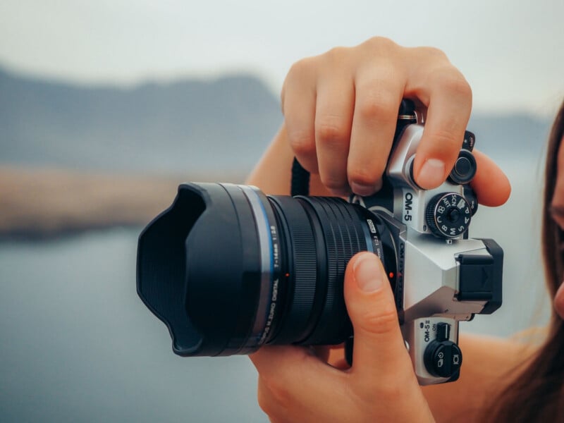 A person adjusts the settings on a silver Olympus OM-5 camera with a wide lens, held outdoors with blurred mountains and water in the background.