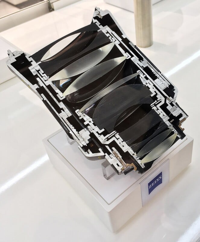 A cross-section of a camera lens on display, showing multiple curved lens elements inside a detailed metal housing, with a “ZEISS” label on the stand beneath it.