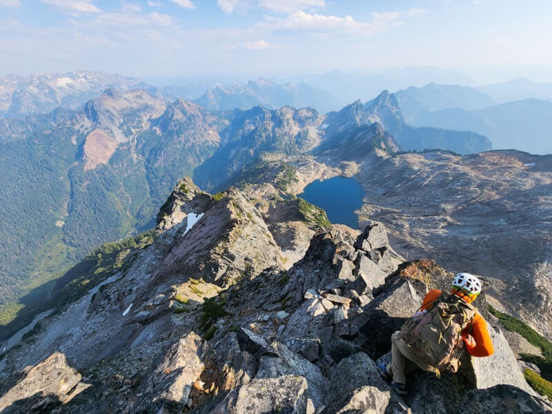A hiker wearing a helmet and backpack rests on rocky terrain atop a mountain ridge, overlooking a scenic view of rugged peaks, a blue alpine lake, and forested valleys under a partly cloudy sky.