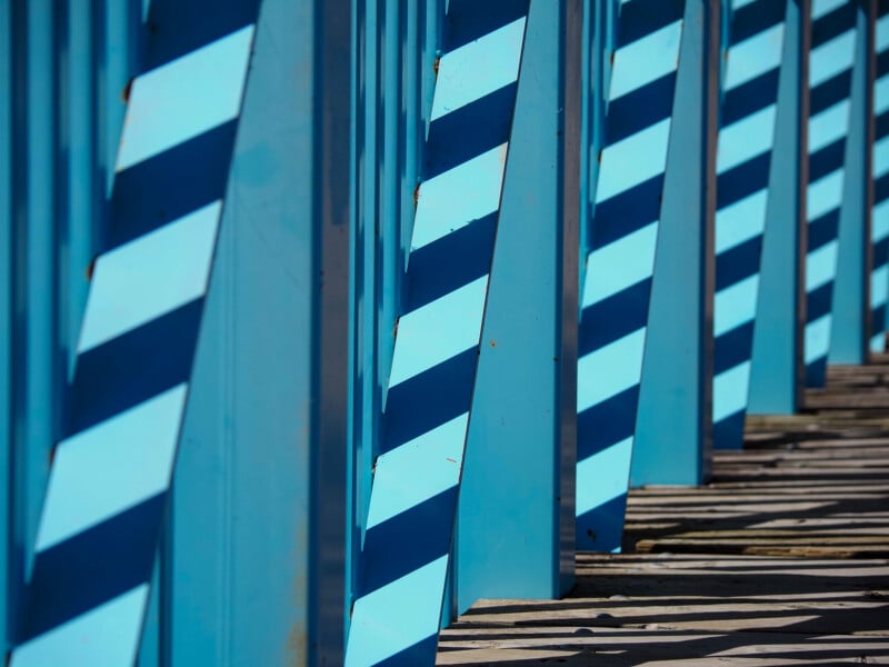 Rows of vertical blue beams cast diagonal shadows on a wooden surface, creating a geometric pattern of alternating light and shadow in bright sunlight.