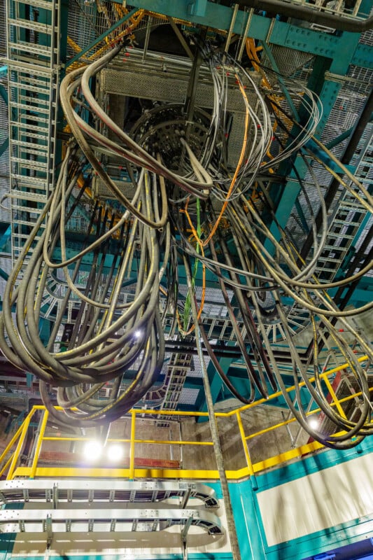 A cluster of thick black cables hangs from the ceiling in an industrial facility with metal platforms, railings, and gridded walkways visible below. Bright lighting highlights the structure and cables.