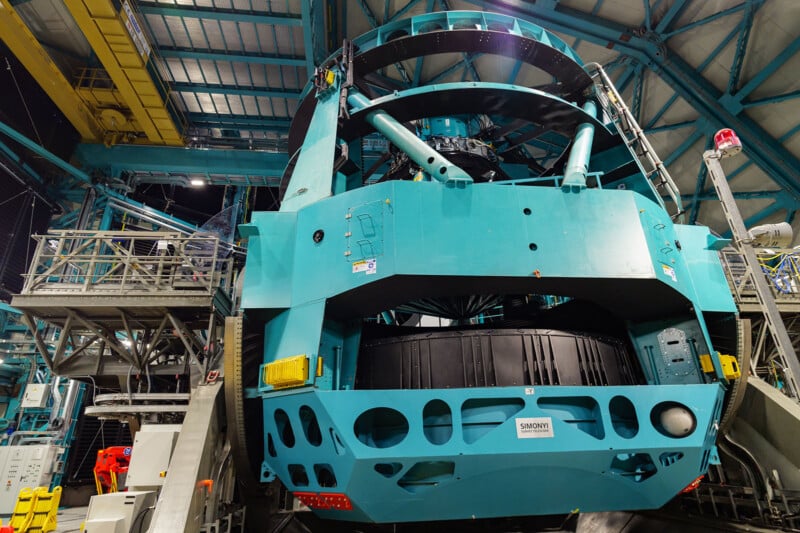 Large blue-green telescope structure inside an observatory, surrounded by metal platforms, beams, and equipment under a high ceiling with industrial lighting.