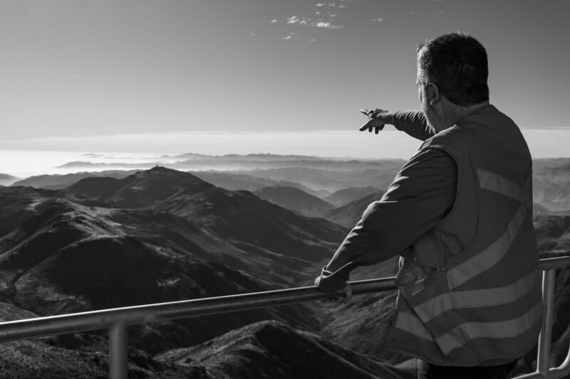 A person in a reflective vest stands at a railing, looking out over a vast mountain landscape and pointing toward the distant horizon under a clear sky.