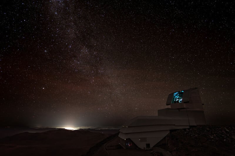 A large observatory sits on a mountaintop under a star-filled night sky, with the Milky Way visible above and distant city lights glowing on the horizon.