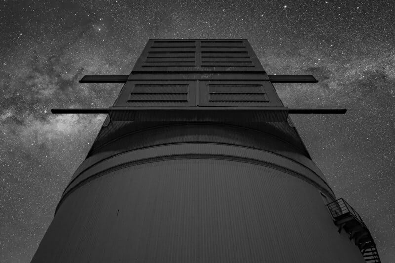 A tall industrial structure shot from below at night, with a dramatic view of the star-filled sky and the Milky Way in the background. The image is in black and white.