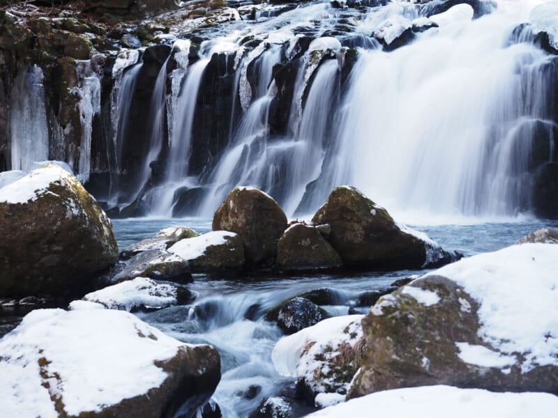 A snowy waterfall cascades over rocks in a winter landscape, with water flowing smoothly between mossy stones and patches of snow covering the ground and rocks.