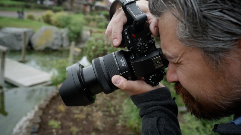 Un hombre barbudo y de cabello gris se acerca una cámara a los ojos y toma fotografías al aire libre cerca de un estanque y un muelle de madera.