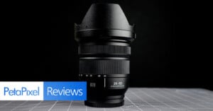A wide-angle camera lens stands upright on a gray surface with grid lines, against a black background. "PetaPixel Reviews" is displayed in blue and white text at the bottom left.