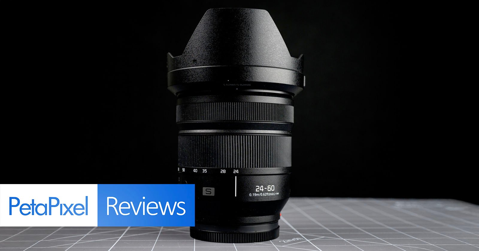 A wide-angle camera lens stands upright on a gray surface with grid lines, against a black background. "PetaPixel Reviews" is displayed in blue and white text at the bottom left.