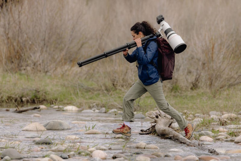 A woman in outdoor gear steps across rocks in a shallow stream, carrying a large camera with a telephoto lens and tripod on her shoulder, surrounded by natural scenery.