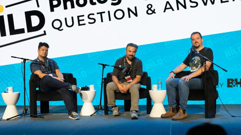 Three men sit on a stage in black chairs with microphones, participating in a photography Q&A session at B&H’s BILD Expo. A large event sign and blue backdrop are visible behind them.