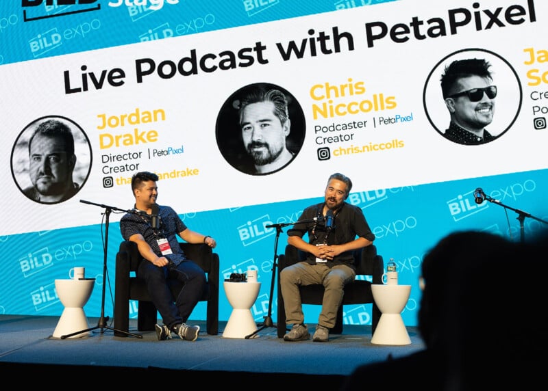 Two men sit on stage with microphones during a live podcast at BILD Expo. Behind them is a large screen displaying their photos, names, and titles: Jordan Drake and Chris Niccolls from PetaPixel.
