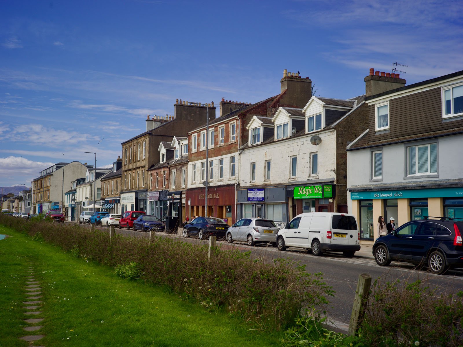 A row of shops and parked cars lines a street in a small town, with multi-story buildings, a grassy area with bushes in the foreground, and a blue sky above. People are walking along the sidewalk.