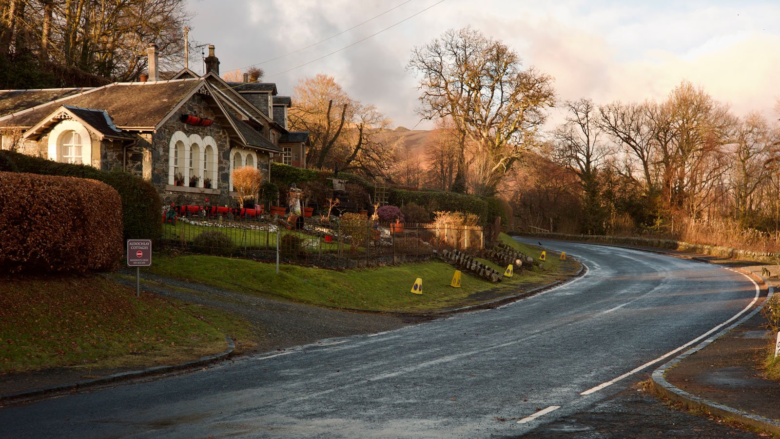 A winding road curves past a cozy stone house with arched windows, red outdoor seating, and landscaped gardens. Leafless trees and soft sunlight suggest a peaceful, late autumn or early winter scene.