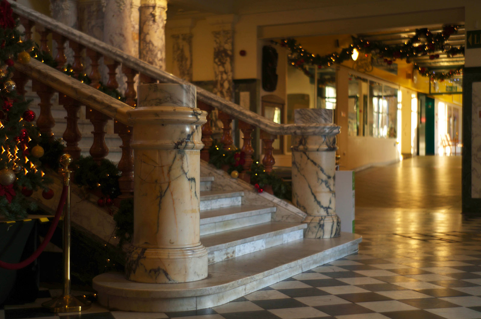 Marble staircase with wooden railings, decorated with Christmas garlands and ornaments. The floor is checkered black and white, and the hallway in the background is warmly lit and festive.