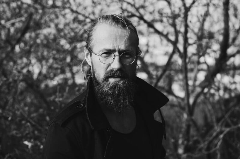 Black and white photo of a bearded man with glasses and slicked-back hair, wearing a dark coat, standing outdoors in front of leafless trees.