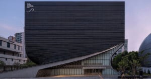 Modern building with a striking, curved facade made of horizontal black slats, partially lifted at one corner to reveal glass windows and an entrance below. Urban surroundings with trees and neighboring structures.