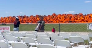 A row of empty white chairs faces a chain-link fence, behind which a massive explosion with bright orange flames occurs. Two people stand near the fence, one holding a large camera lens, both watching the blast.
