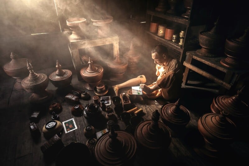 A monk in orange robes sits on a wooden floor, reading a book in soft sunlight surrounded by traditional pots and jars inside a dim, rustic room.
