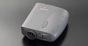 A gray Apple QuickTake 100 digital camera with a rounded body, lens on the front, and Apple logo on top, placed on a sleek dark surface.