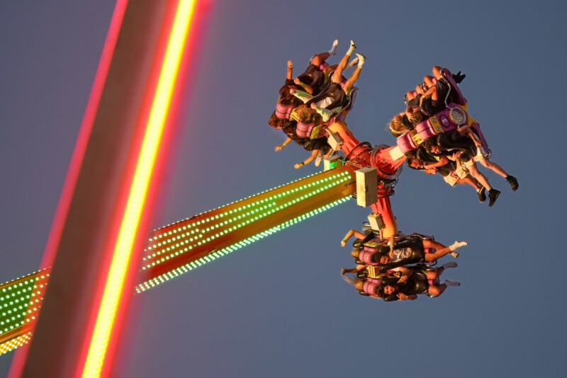 People are strapped into seats on an amusement park ride spinning high in the air at dusk, with colorful lights illuminating the ride against a clear evening sky.