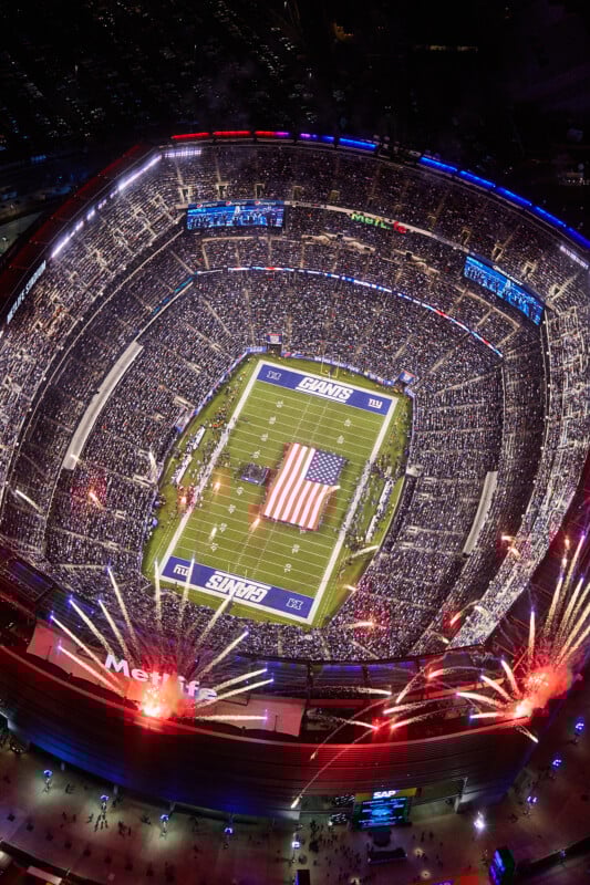 Aerial view of a packed football stadium at night, with an American flag covering the field, surrounded by players and staff. Fireworks burst near the stadium entrance as fans fill the seats, and bright lights illuminate the scene.