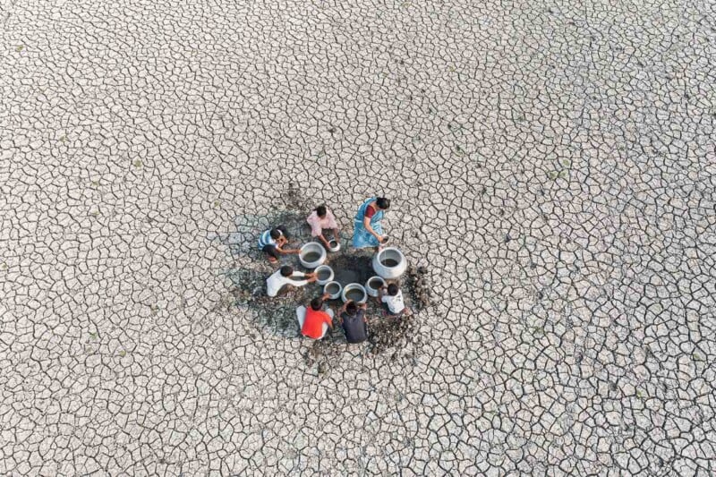 A group of people gathers around empty containers by a small well on cracked, parched earth, highlighting water scarcity in a drought-affected area.