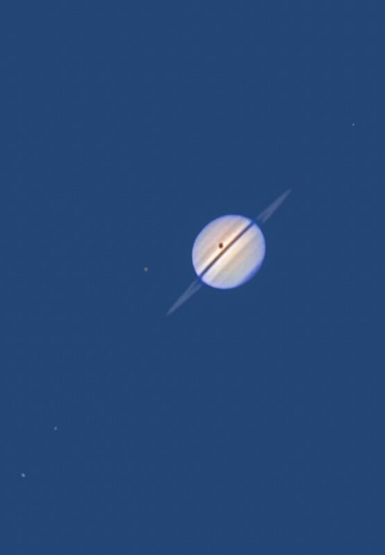 A clear image of Saturn with its rings visible, showing a dark shadow across the planet. Several small bright dots, likely moons or stars, appear around Saturn against a deep blue background.