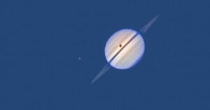 A close-up view of Saturn with its rings clearly visible, showing a dark shadow cast on the planet. A small bright moon is seen nearby against a deep blue background.