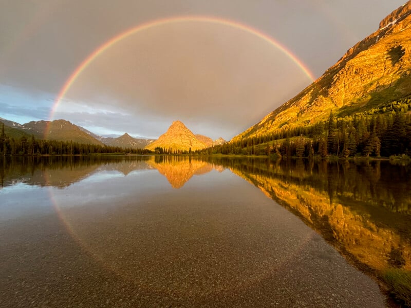 A vivid rainbow arches over a mountain reflected in a calm lake, surrounded by trees and sunlit rocky slopes under a partly cloudy sky.