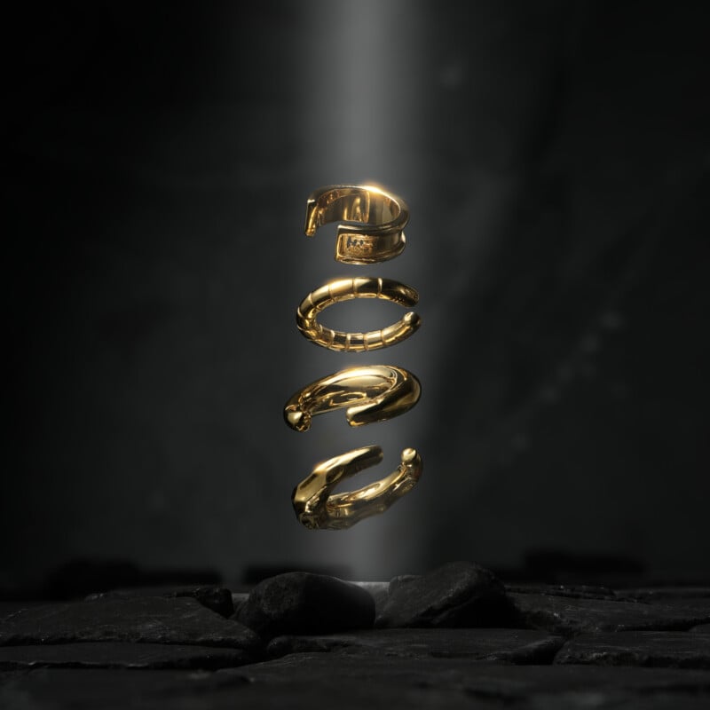 Four abstract, gold metallic shapes float vertically above black stones, illuminated by a beam of light against a dark, textured background.