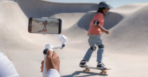 A person holds a smartphone mounted on a gimbal, recording a child wearing a helmet and pads skateboarding in a concrete skatepark.