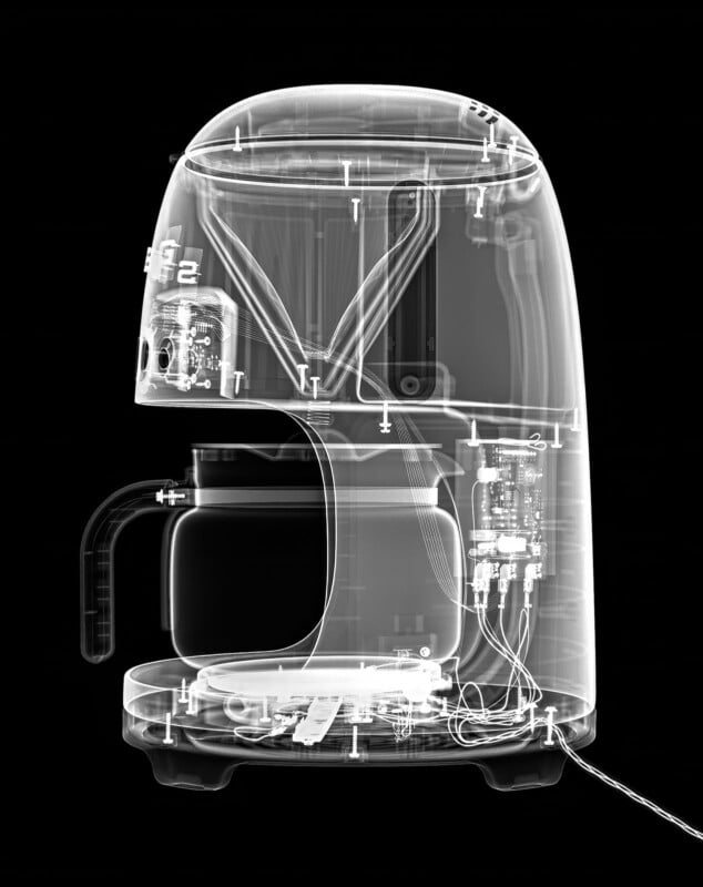 X-ray image of a coffee maker showing its internal components, including wiring, circuit boards, water reservoir, carafe, and structural parts, all visible within a transparent outer shell against a black background.