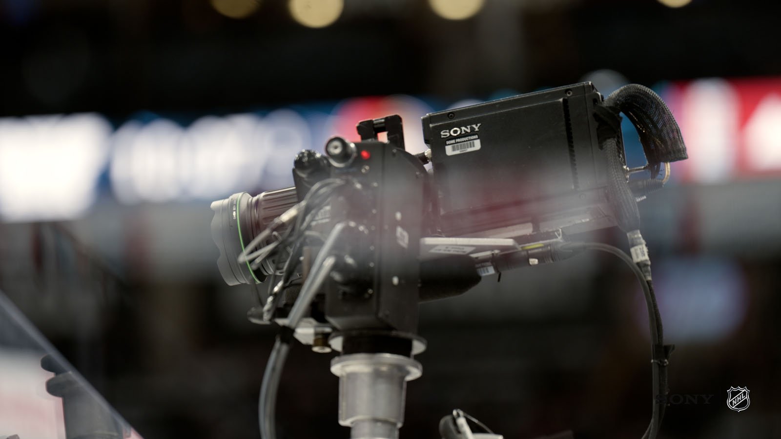 A close-up of a Sony broadcast camera mounted on a stand at an indoor sports venue, with blurred lights and spectators in the background.