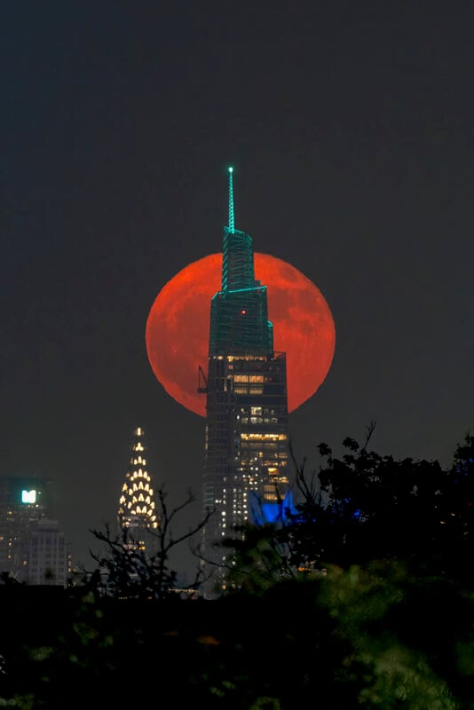 A large, glowing red moon rises behind a tall city skyscraper at night, with other illuminated buildings and dark tree silhouettes in the foreground.