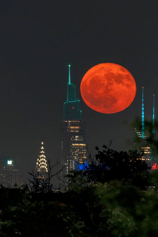 A large, bright red moon rises above a city skyline at night, with tall illuminated skyscrapers and trees in the foreground.