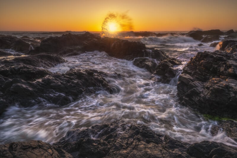 Waves crashing against dark rocks at sunset, with water splashing high and glowing in the golden light. The ocean is turbulent, and the sky is clear and orange near the horizon.