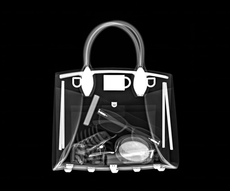 X-ray image of a handbag showing contents including a mug, sunglasses, keys, a tube, a makeup brush, and what appears to be a wallet or small pouch, all displayed in shades of white and gray on a black background.