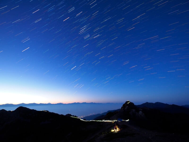 Long-exposure photo of a mountain landscape at night, with streaks of stars moving across the sky and a faint glow on the horizon. A campsite with a glowing tent is visible at the base of the mountain.