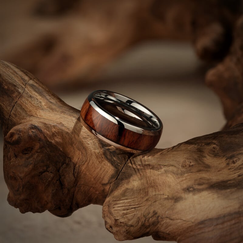 A polished ring with a wood inlay and metallic edges rests on a textured piece of driftwood, highlighting the contrast between the smooth band and the rugged natural surface.