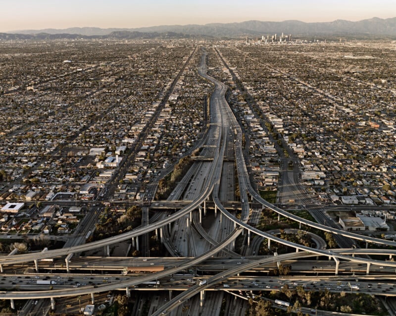 Aerial view of a sprawling freeway interchange in Los Angeles, surrounded by densely packed houses and buildings, with downtown skyscrapers and mountains visible in the distance under a hazy sky.