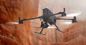 A black quadcopter drone with a mounted camera flying in a narrow, sunlit canyon with reddish-brown rock walls. The drone's propellers are blurred, indicating it is in motion.