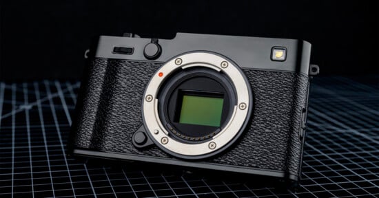 A black digital camera body without a lens is displayed on a grid-patterned surface, showing its sensor and textured grip, with a dark background behind it.
