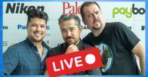Three men pose in front of a wall with sponsor logos. One man puts his arm around another; all are smiling or making expressive faces. A red "LIVE" icon with a white dot appears over the lower center of the image.